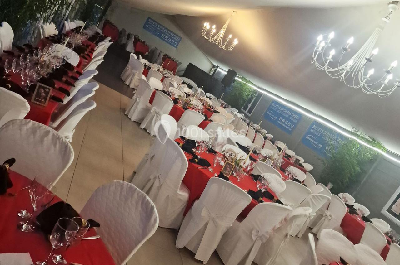 Salle de réception décorée avec des tables rondes, nappes rouges et blanches, chaises couvertes et lustres suspendus.