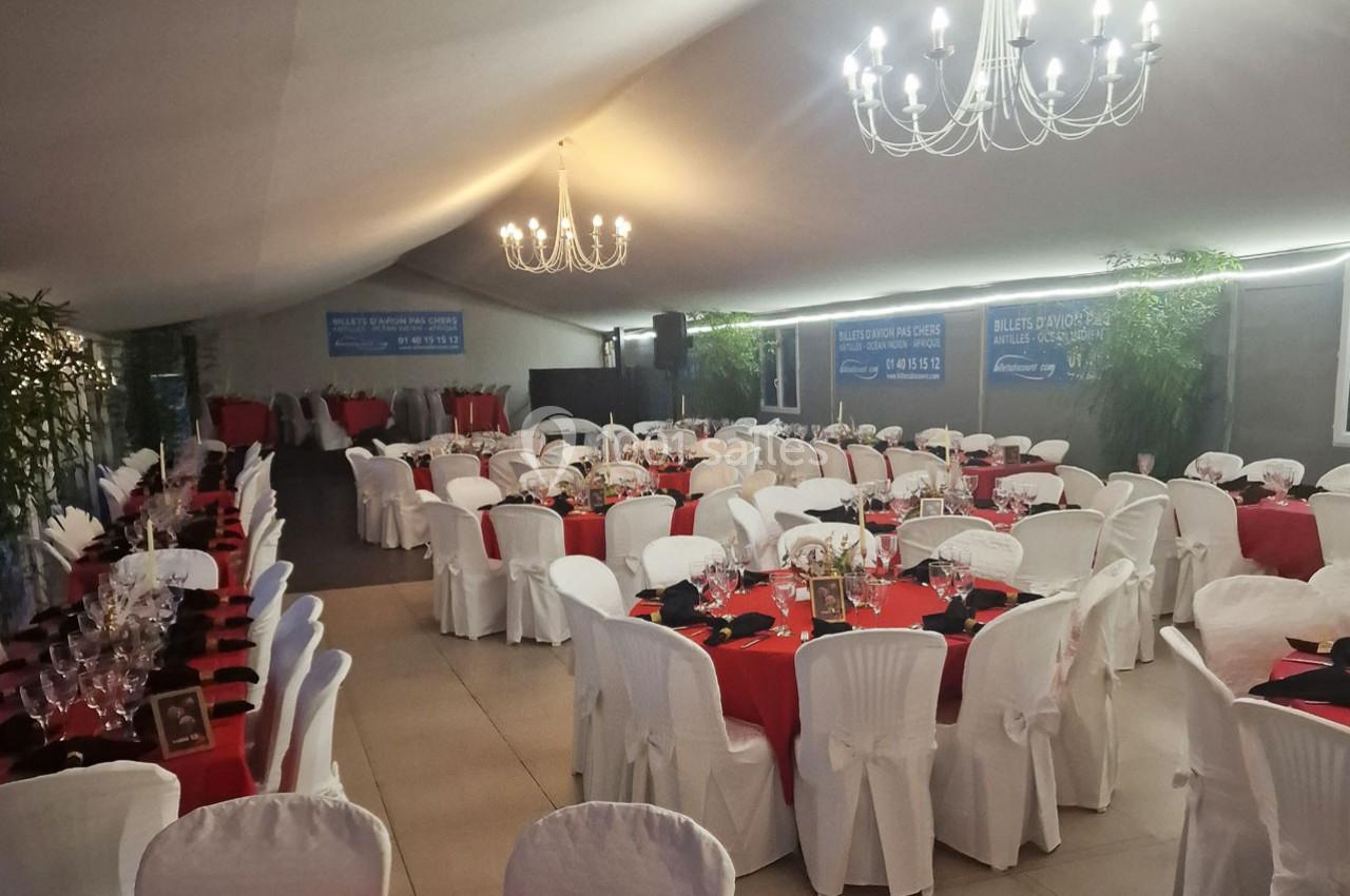 Salle de réception décorée avec des tables rondes, nappes rouges et blanches, chaises couvertes et lustres suspendus.