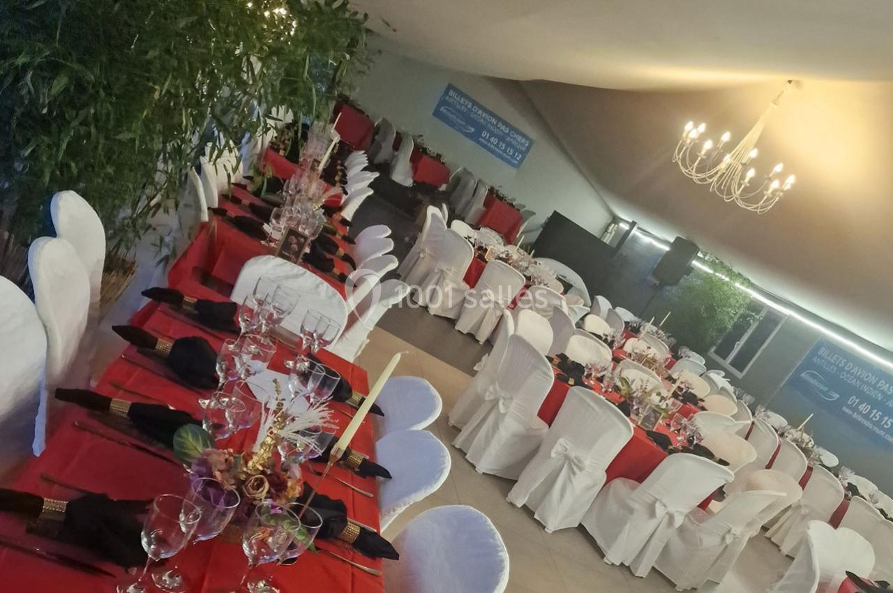 Salle de réception décorée avec des tables dressées, nappes rouges, chaises blanches et un lustre suspendu.