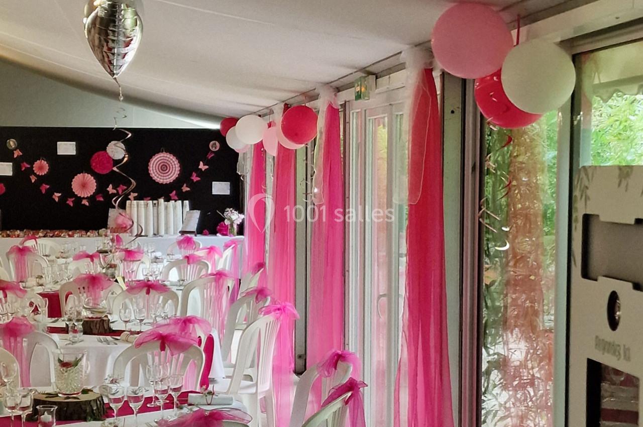 Salle décorée pour une réception avec nappes blanches, chaises ornées de rubans roses et ballons suspendus.