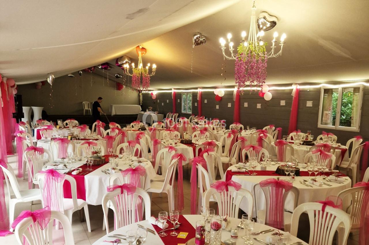 Salle de réception décorée en rose et blanc avec des tables rondes dressées et des chaises ornées de nœuds roses.