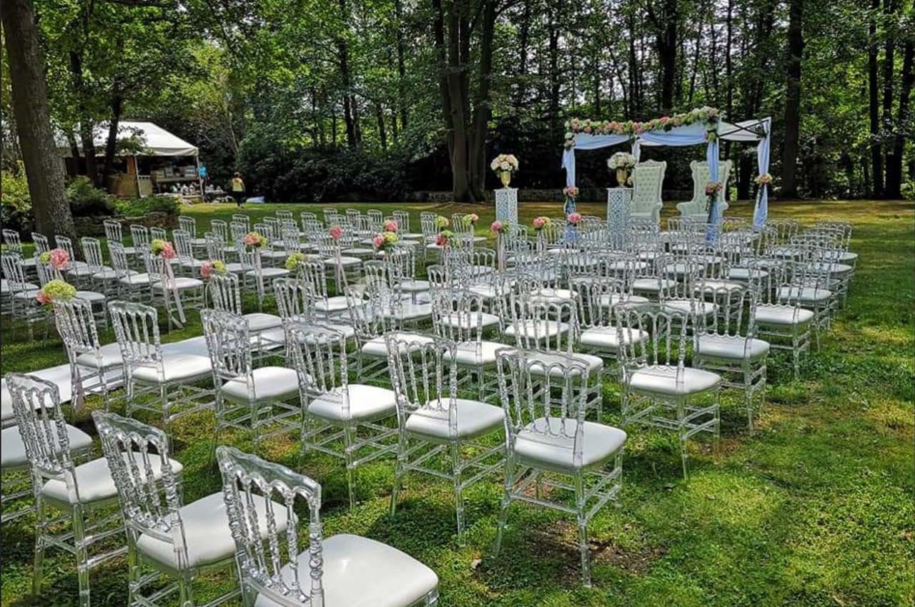 Chaises transparentes alignées en extérieur devant une arche décorée pour une cérémonie dans un jardin boisé.