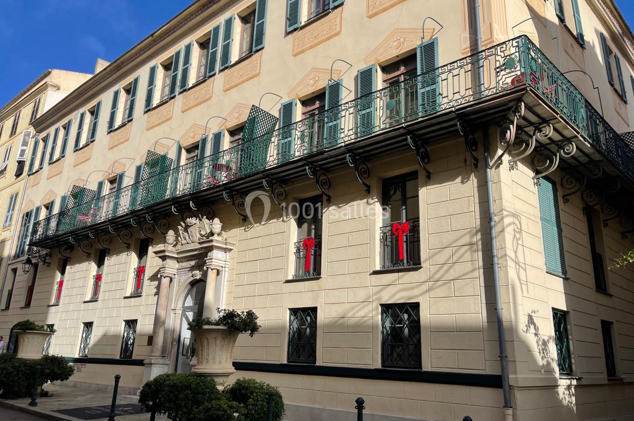 Façade d'un bâtiment historique avec balcons en fer forgé, volets verts et décorations rouges sur les fenêtres.