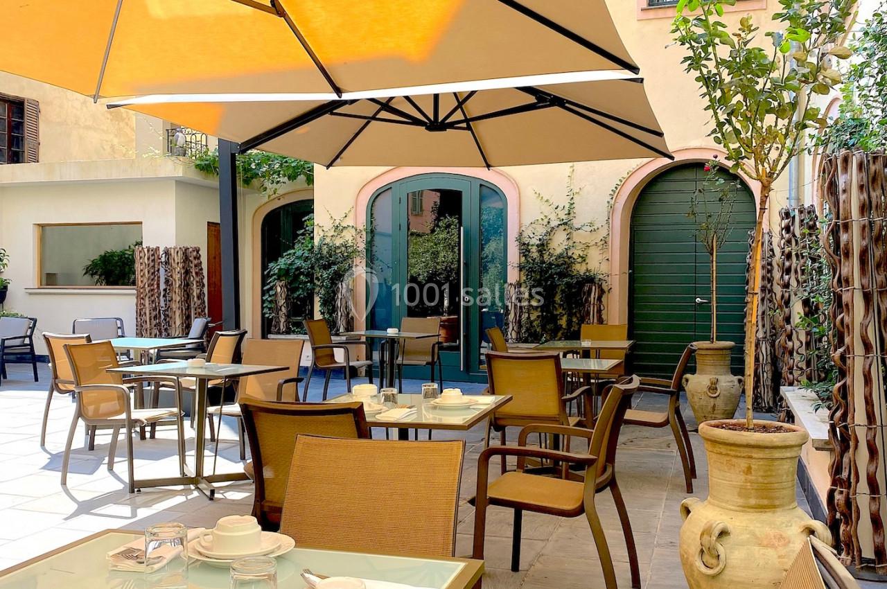 Terrasse ensoleillée avec tables, chaises, parasols et plantes en pot dans une cour pavée.