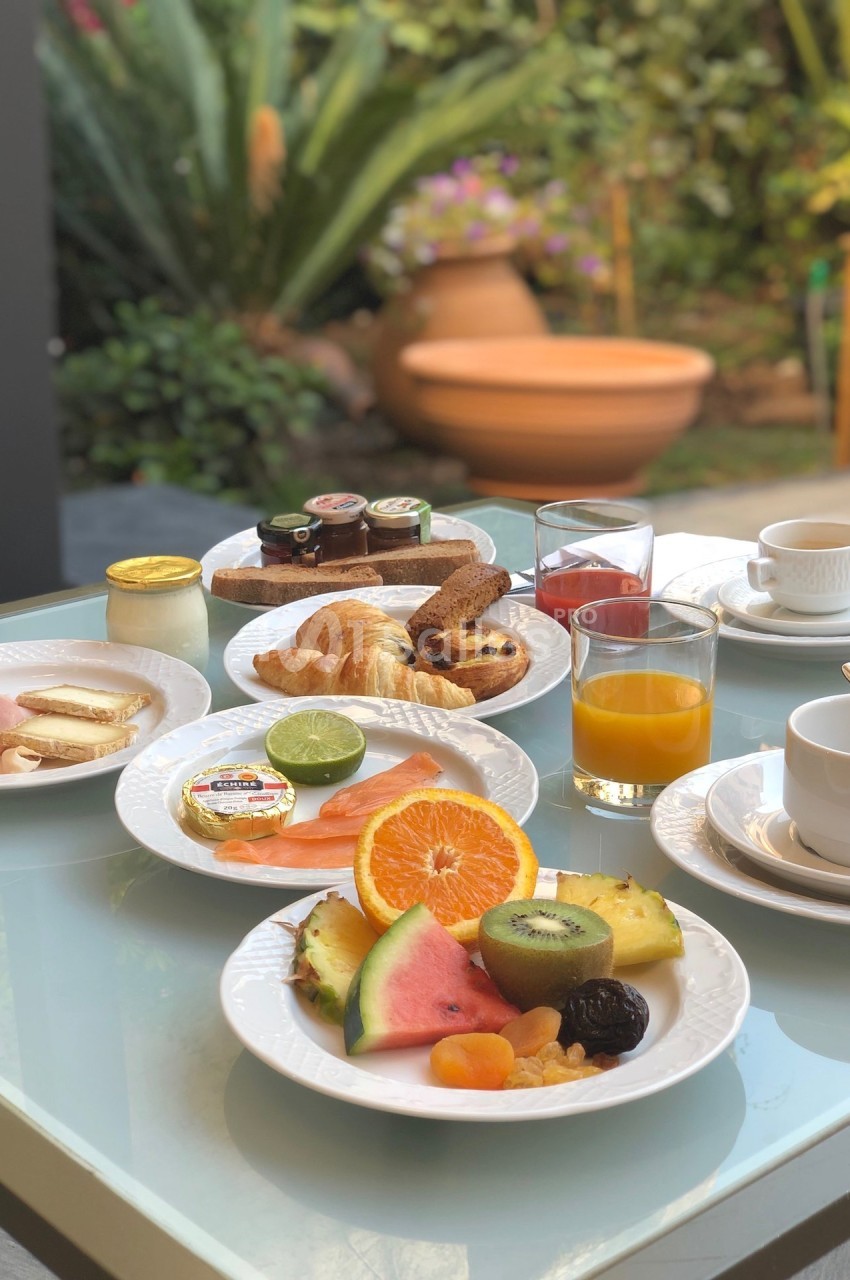 Petit-déjeuner varié avec fruits, viennoiseries, saumon, charcuterie, confitures et boissons sur une table en extérieur.