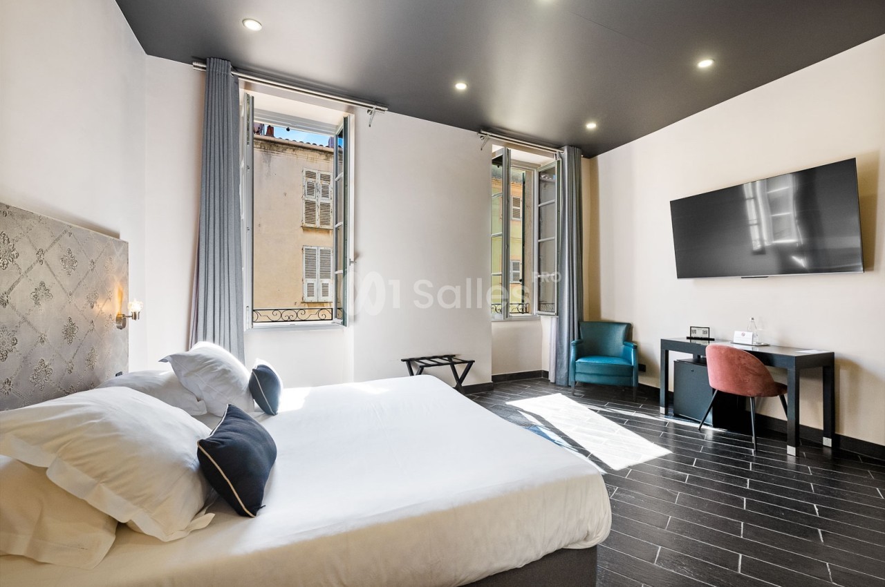 Chambre lumineuse avec lit double, bureau, fauteuil, télévision murale et fenêtres ouvertes sur des bâtiments voisins.