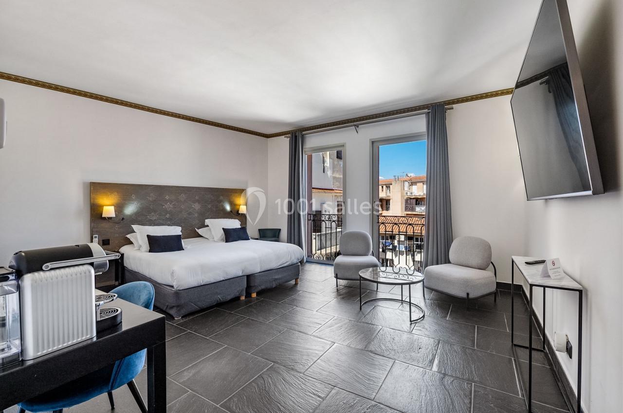 Chambre lumineuse avec lit double, coin salon, bureau, balcon et vue sur des bâtiments voisins sous un ciel bleu.