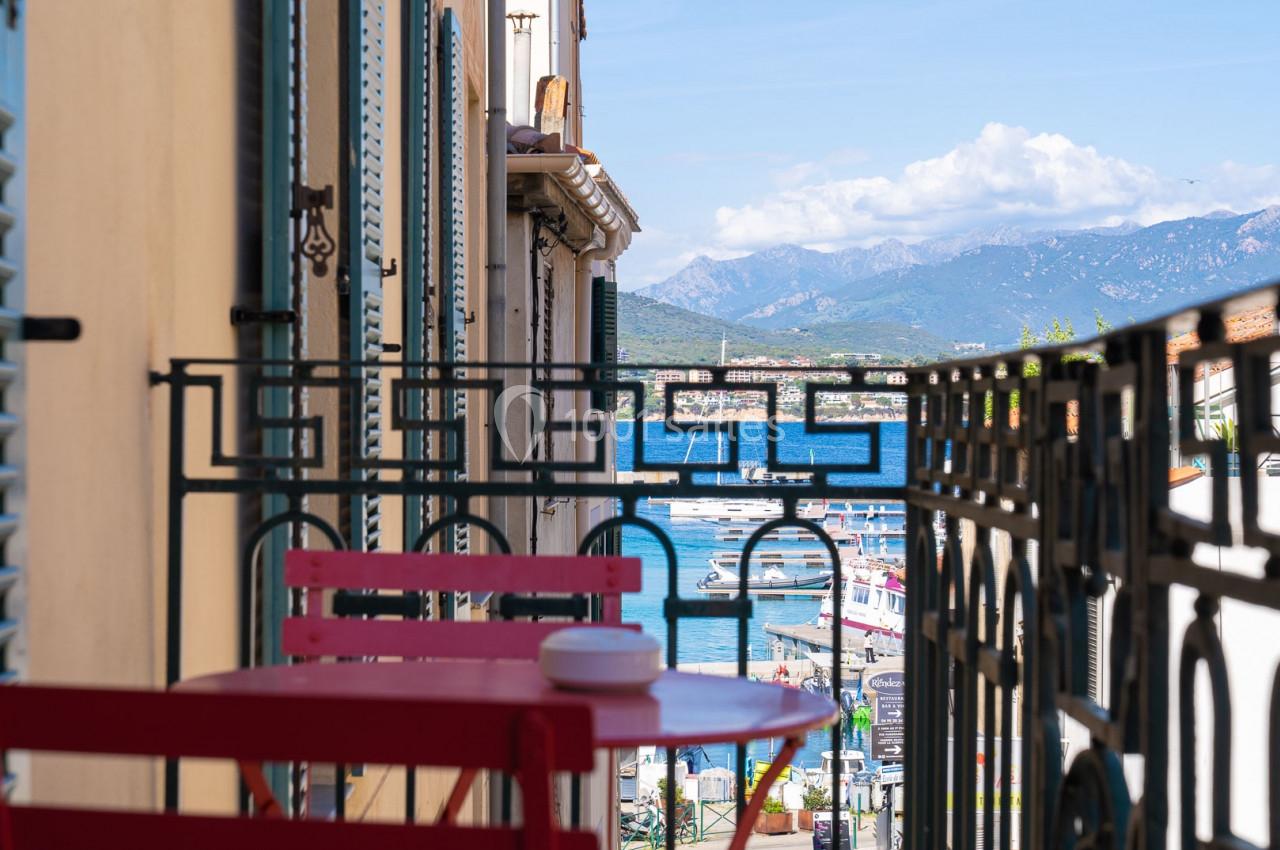 Vue depuis un balcon avec une table rouge, donnant sur un port, des bâtiments et des montagnes en arrière-plan.
