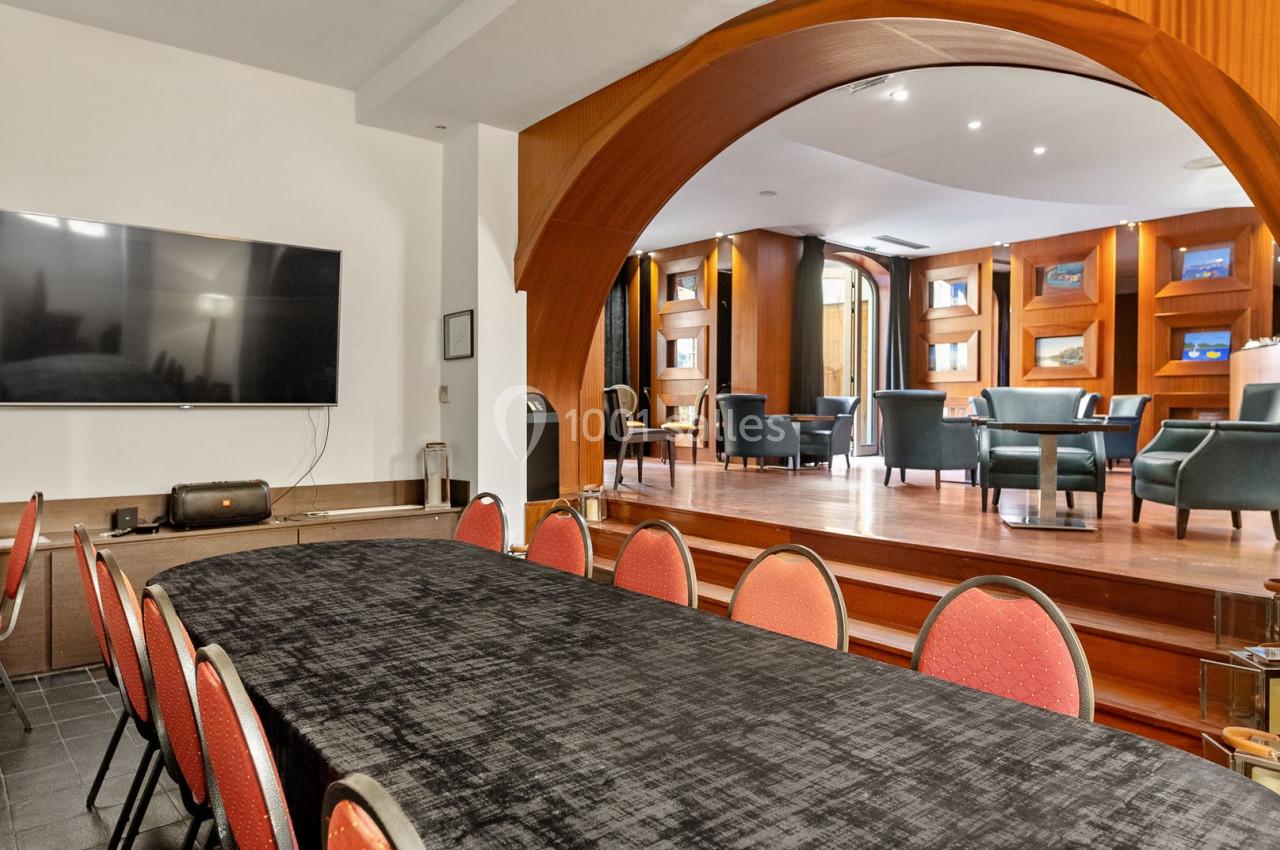 Salle avec une grande table recouverte d'une nappe noire, des chaises rouges et un espace lounge en arrière-plan.