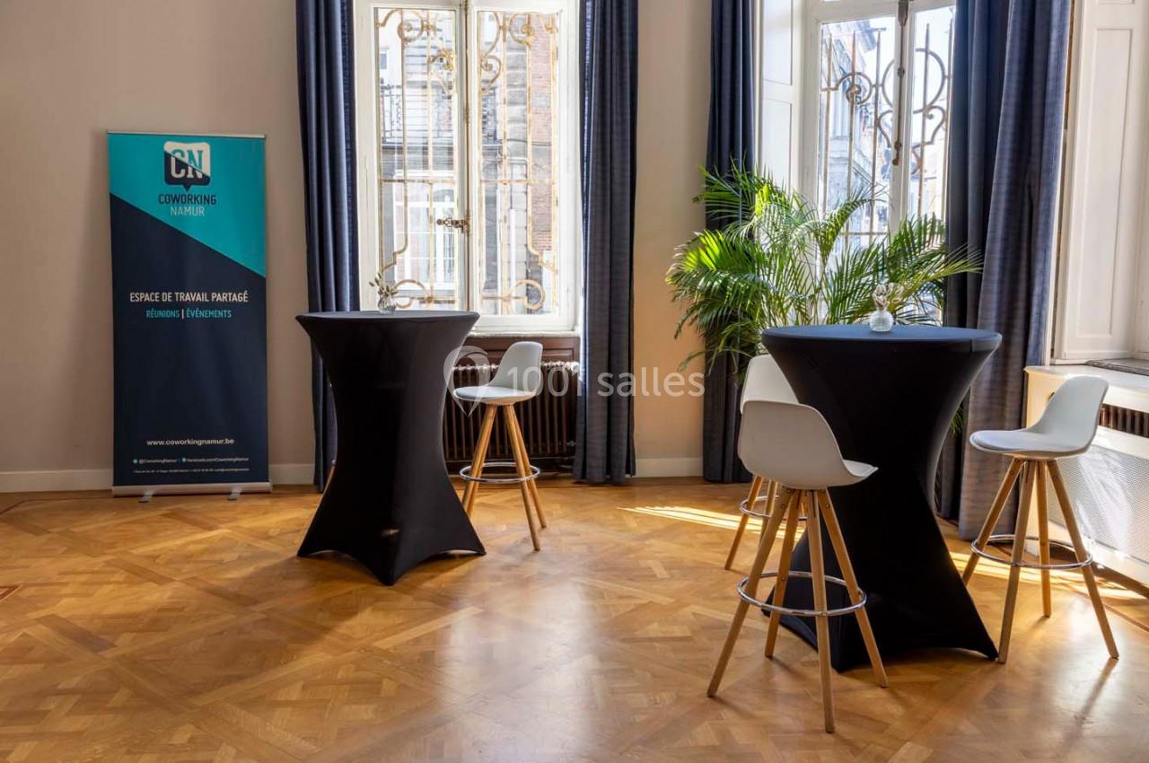 Salle lumineuse avec tables hautes, chaises blanches, plantes et une bannière d'espace de travail partagé près des fenêtres.
