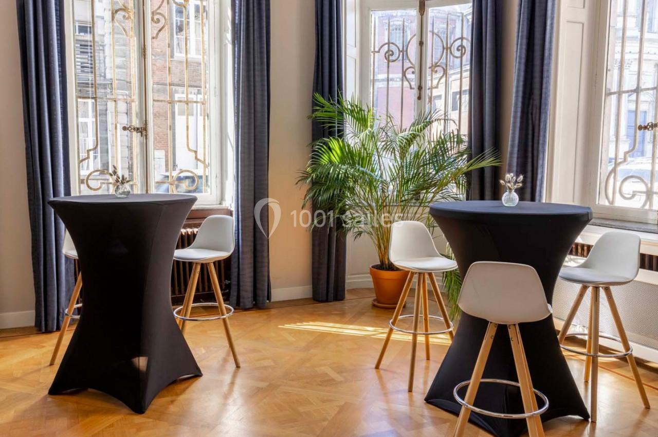 Salle lumineuse avec parquet, tables hautes noires, chaises blanches et plante verte près des fenêtres.