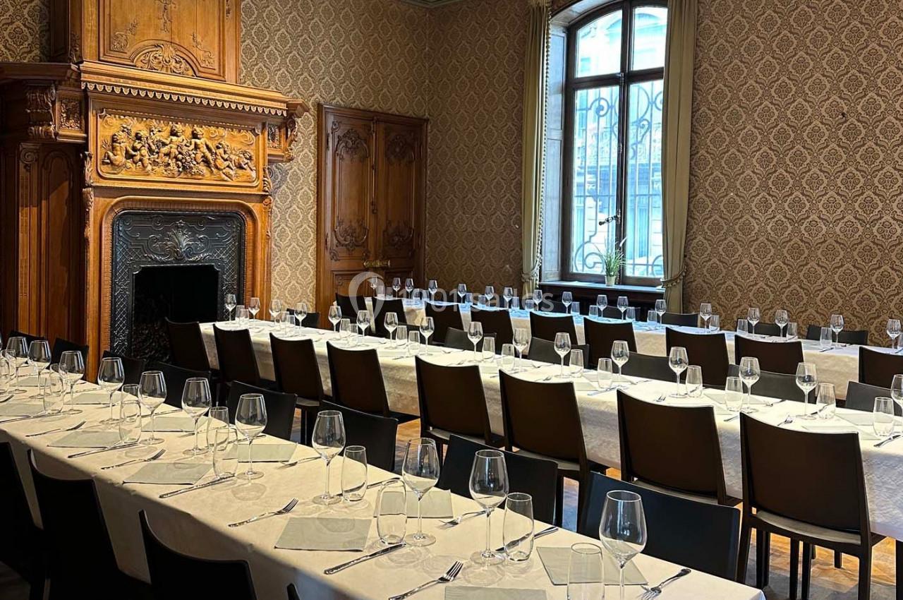 Salle élégante avec de longues tables dressées, nappes blanches, verres et couverts, devant une cheminée sculptée.