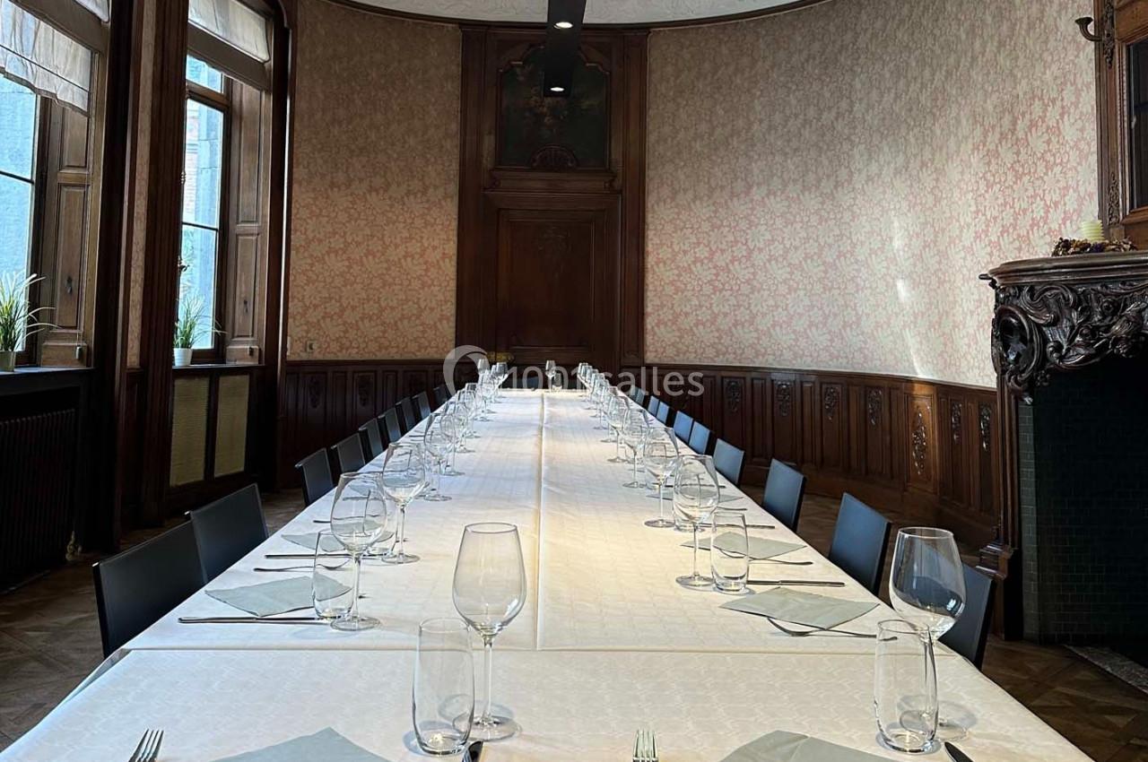 Grande table dressée avec nappes blanches, verres et serviettes, dans une salle élégante aux boiseries et hauts plafonds.