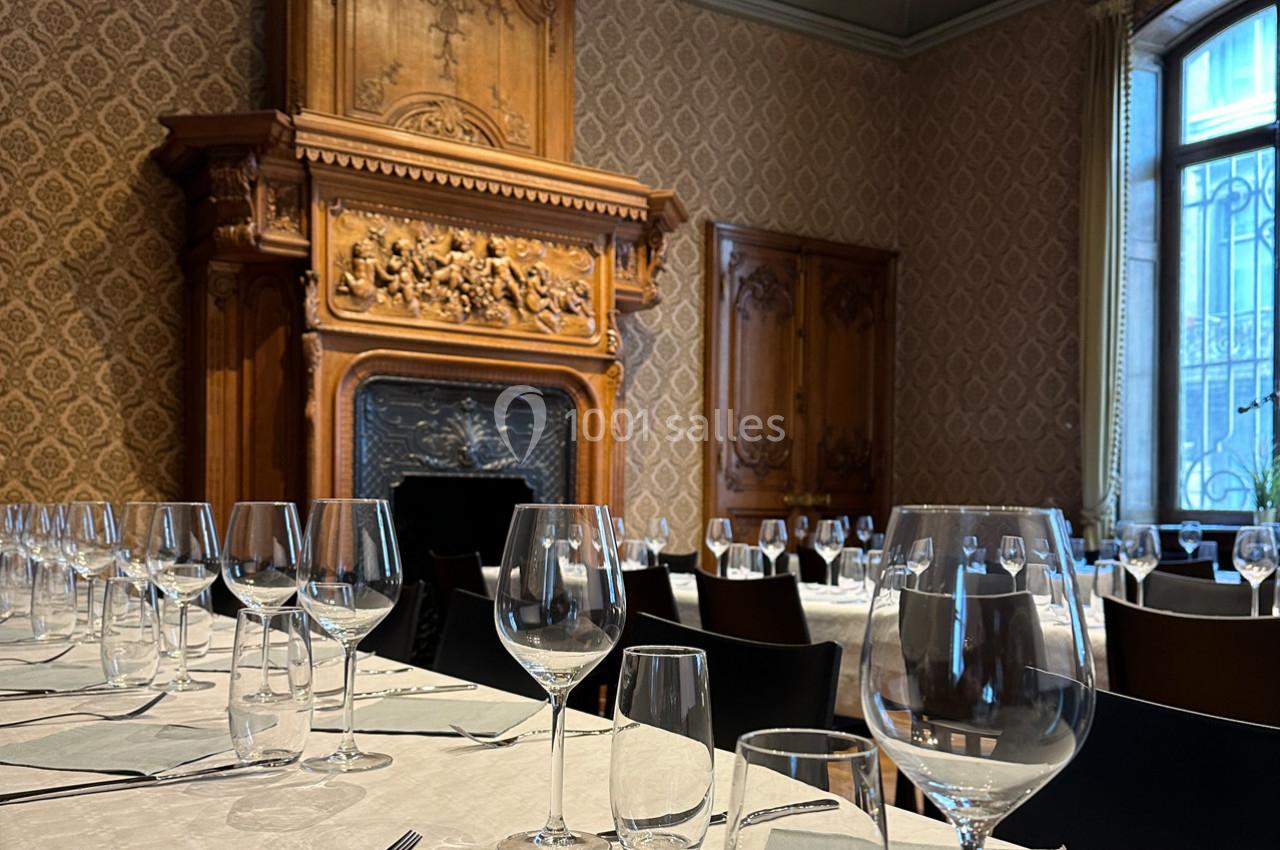 Salle à manger élégante avec cheminée sculptée en bois, tables dressées avec nappes blanches et verres alignés.