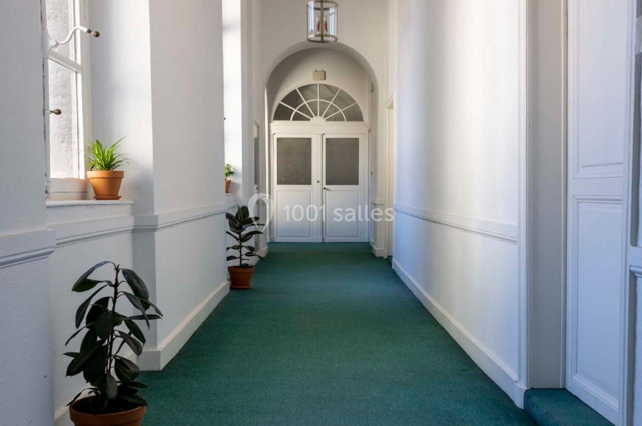 Couloir lumineux avec moquette verte, plantes en pot et porte double vitrée au fond.