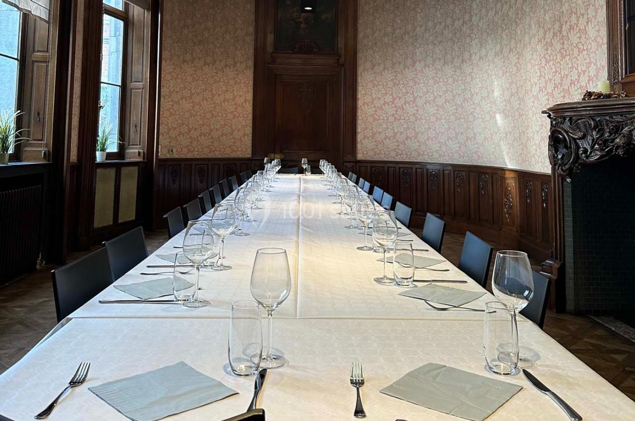 Grande table rectangulaire dressée avec des verres, couverts et serviettes dans une salle élégante aux boiseries anciennes.