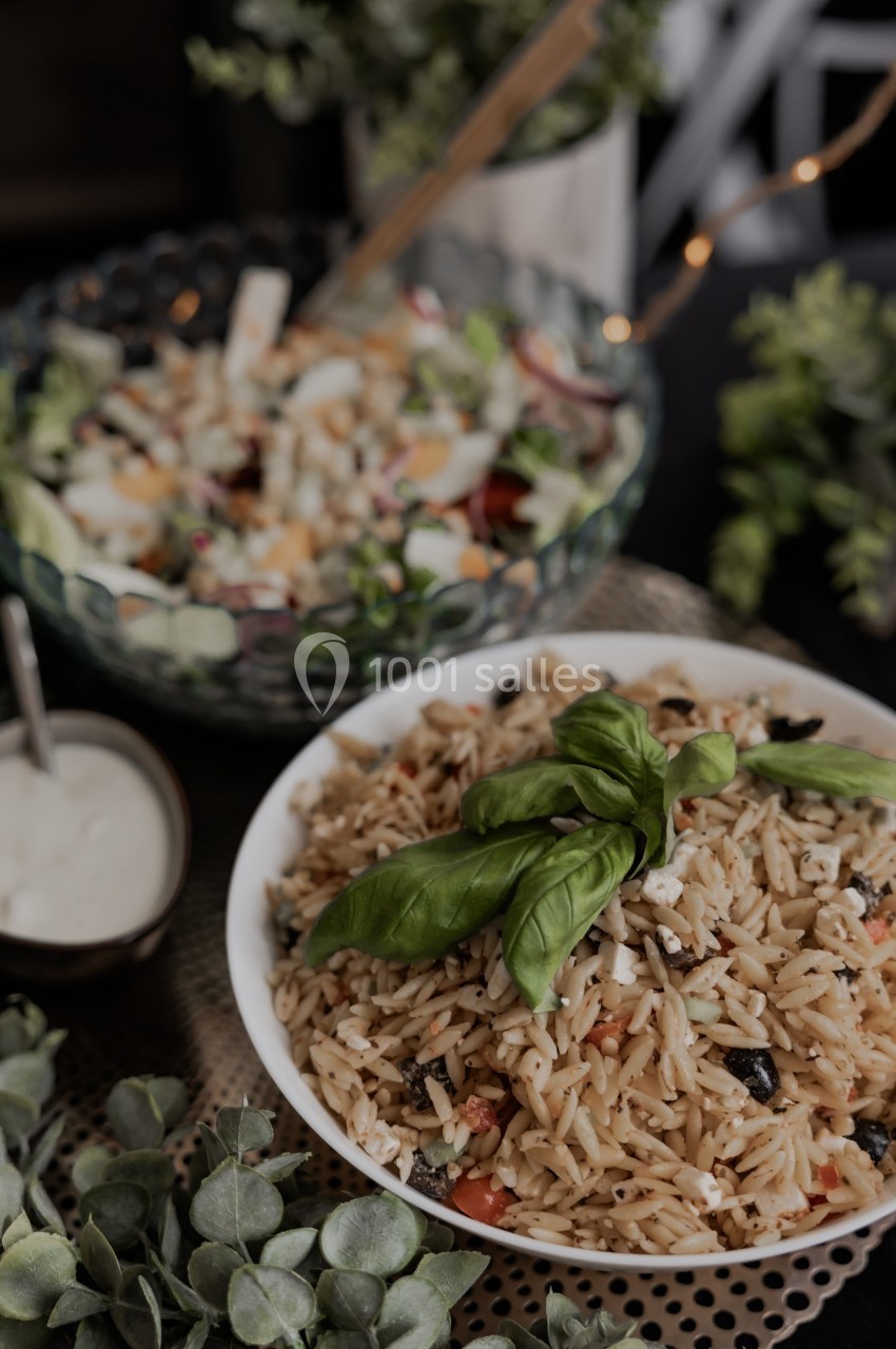 Assiette de salade d'orzo garnie de feuilles de basilic, avec en arrière-plan une salade composée et une sauce.
