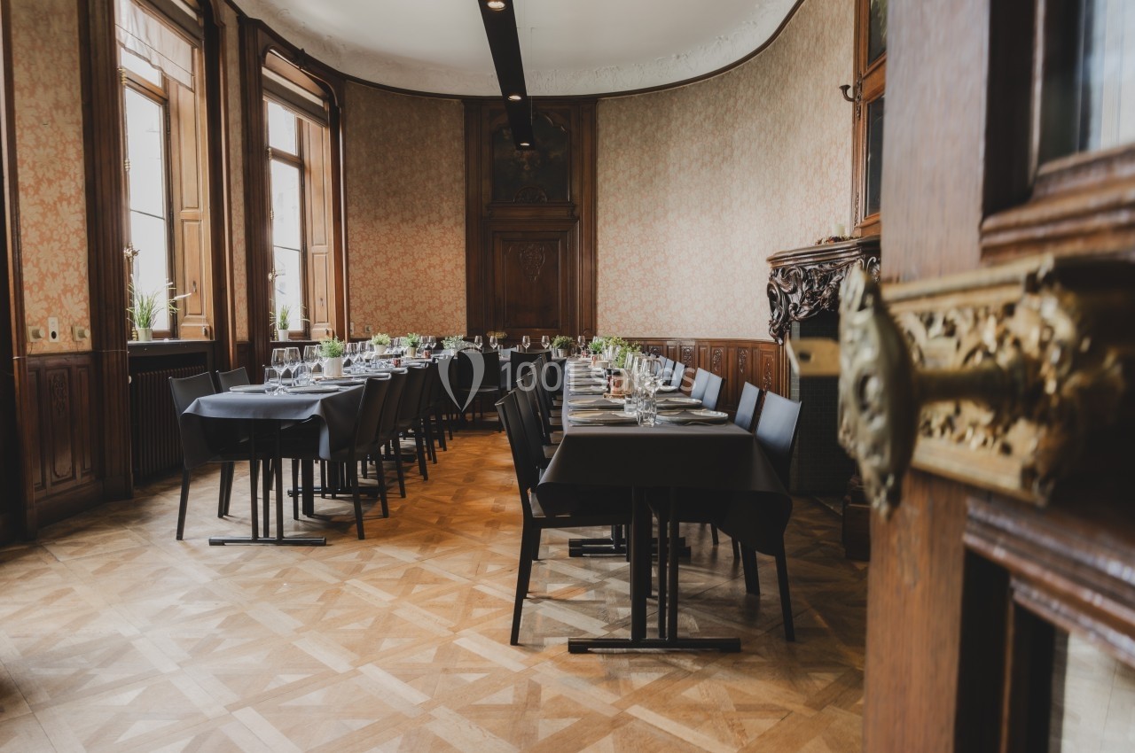 Salle élégante avec tables dressées, chaises noires et grandes fenêtres laissant entrer la lumière naturelle.