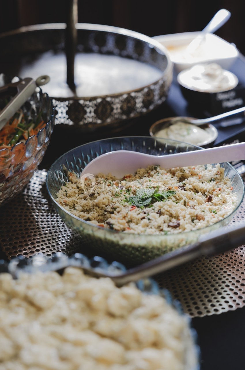 Salade de taboulé servie dans un bol en verre, accompagnée d'ustensiles et d'autres plats en arrière-plan.