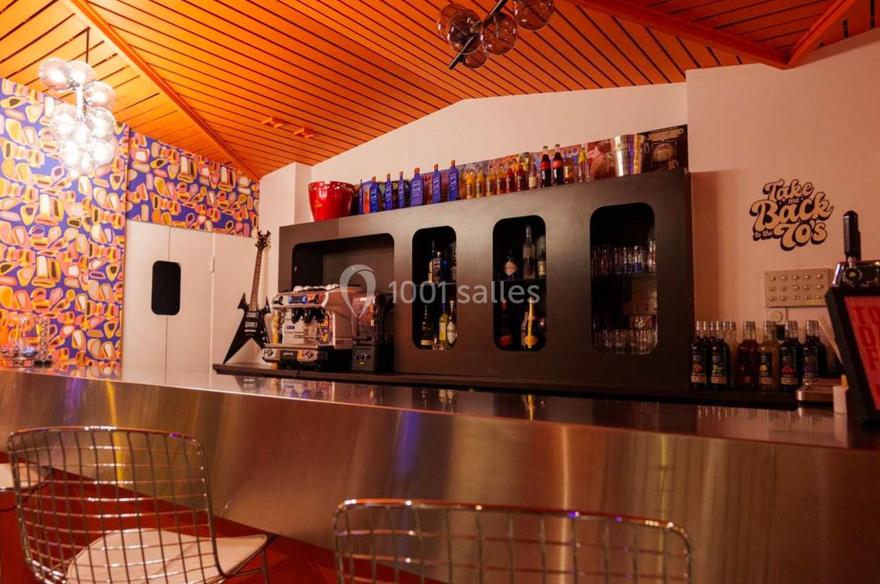 Intérieur d'un bar rétro avec comptoir métallique, étagères de bouteilles et décoration colorée sur les murs.