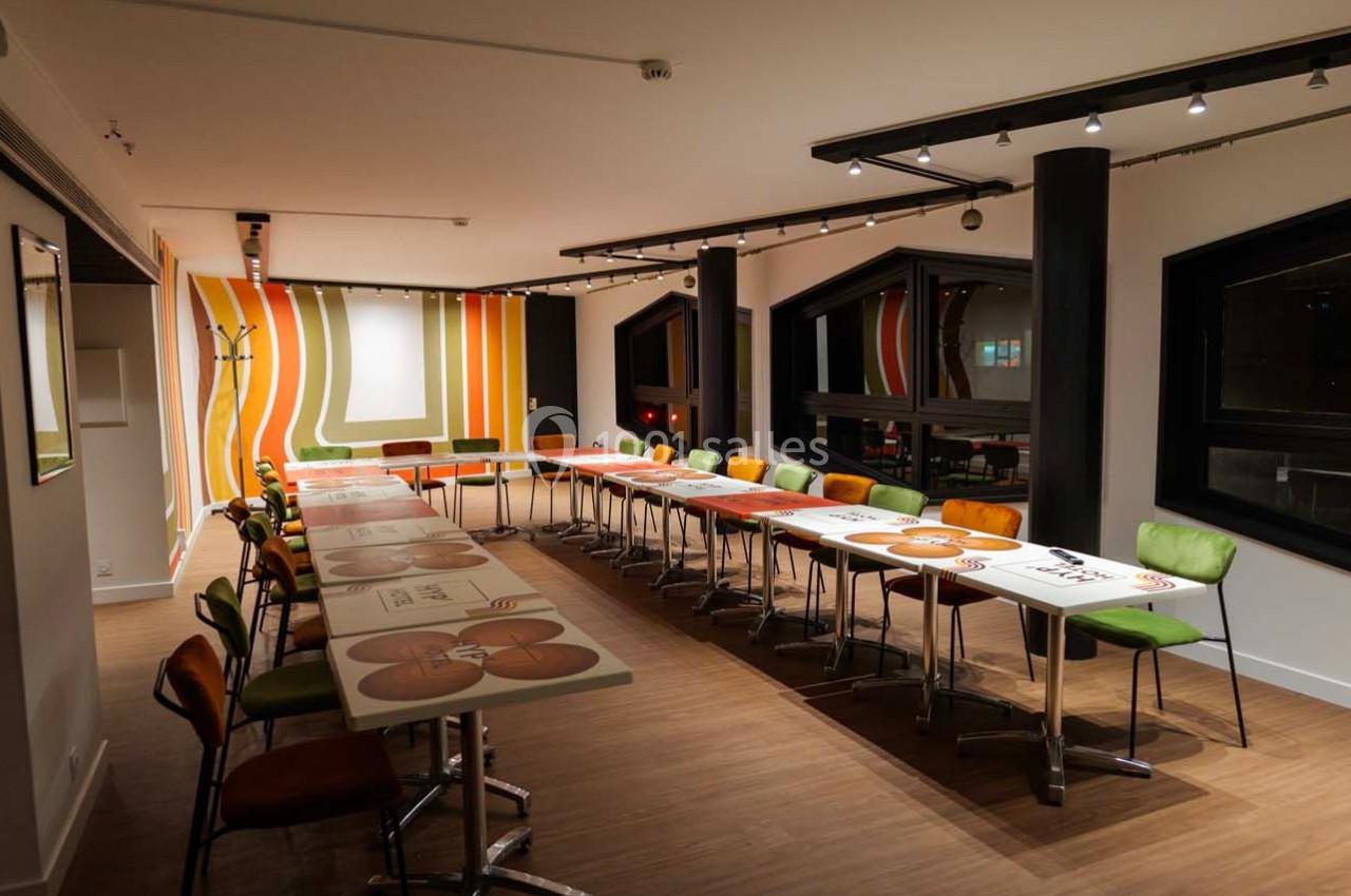 Salle de réunion moderne avec tables disposées en U, chaises colorées et décoration murale aux motifs géométriques.