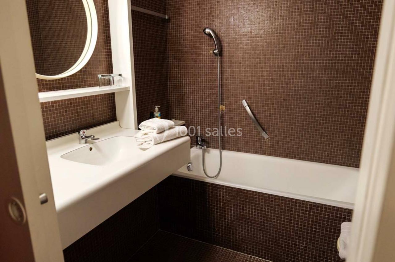 Salle de bain avec carrelage marron, lavabo blanc, miroir rond, baignoire avec douchette et serviettes pliées.