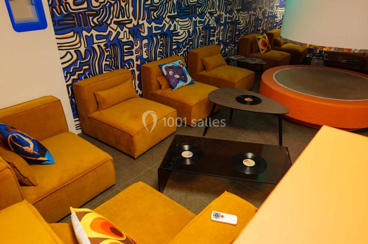 Salon avec fauteuils jaunes, table basse noire avec des vinyles, mur décoré de motifs abstraits bleus et blancs.