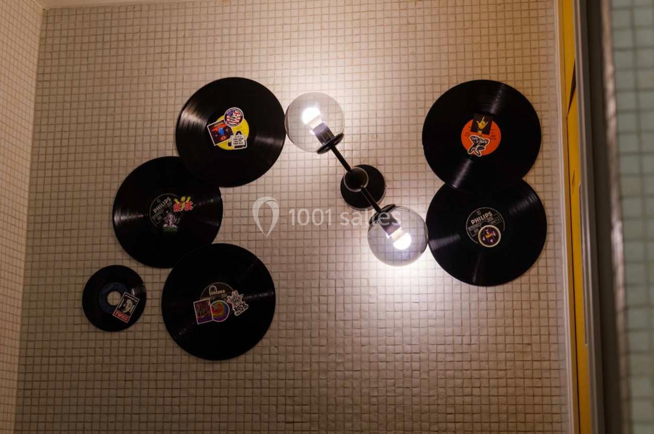 Disques vinyles décoratifs fixés sur un mur carrelé beige, éclairés par une applique murale noire à deux ampoules.