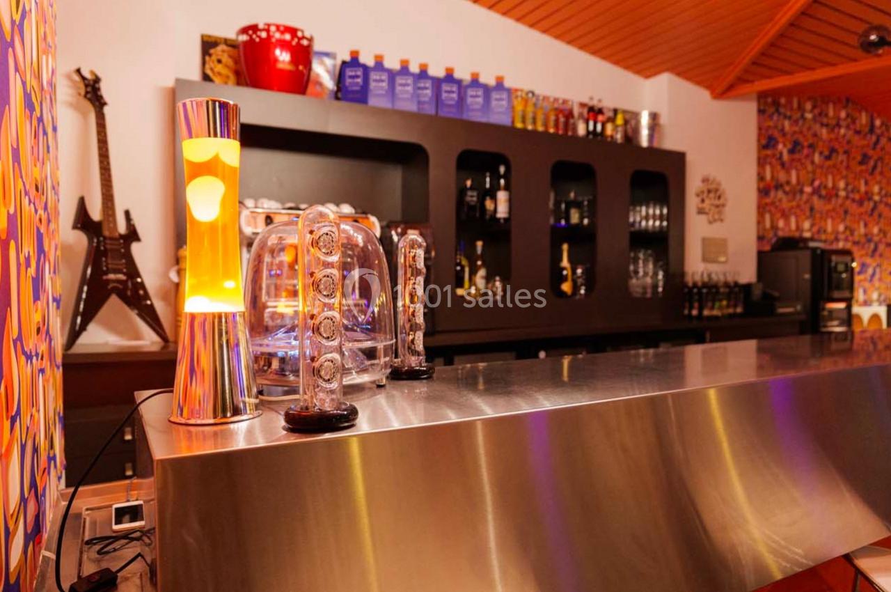 Bar avec comptoir en métal, lampe à lave, décorations colorées et étagères contenant des bouteilles à l’arrière.