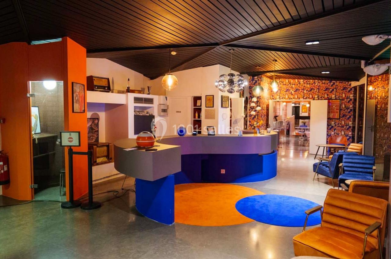 Espace d'accueil moderne avec comptoir bleu, fauteuils colorés et décoration rétro sous un plafond en bois sombre.