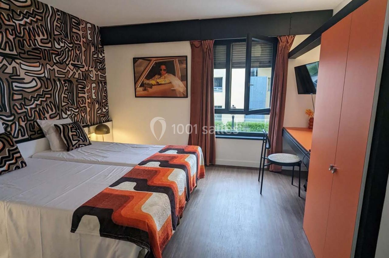 Chambre avec deux lits simples, décorée de motifs géométriques, avec une fenêtre, un bureau et une armoire orange.