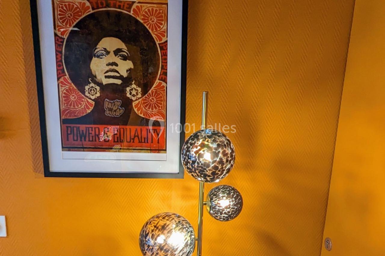 Affiche encadrée sur un mur jaune avec une lampe à trois globes lumineux au premier plan.