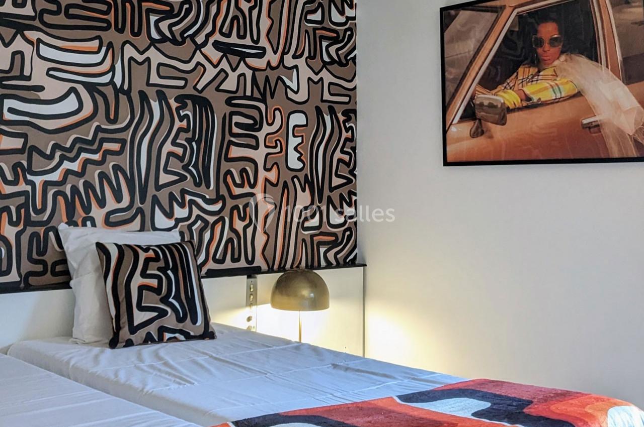 Lit simple avec linge blanc, mur décoré d’un motif abstrait, lampe dorée et photo encadrée d’une femme dans une voiture.