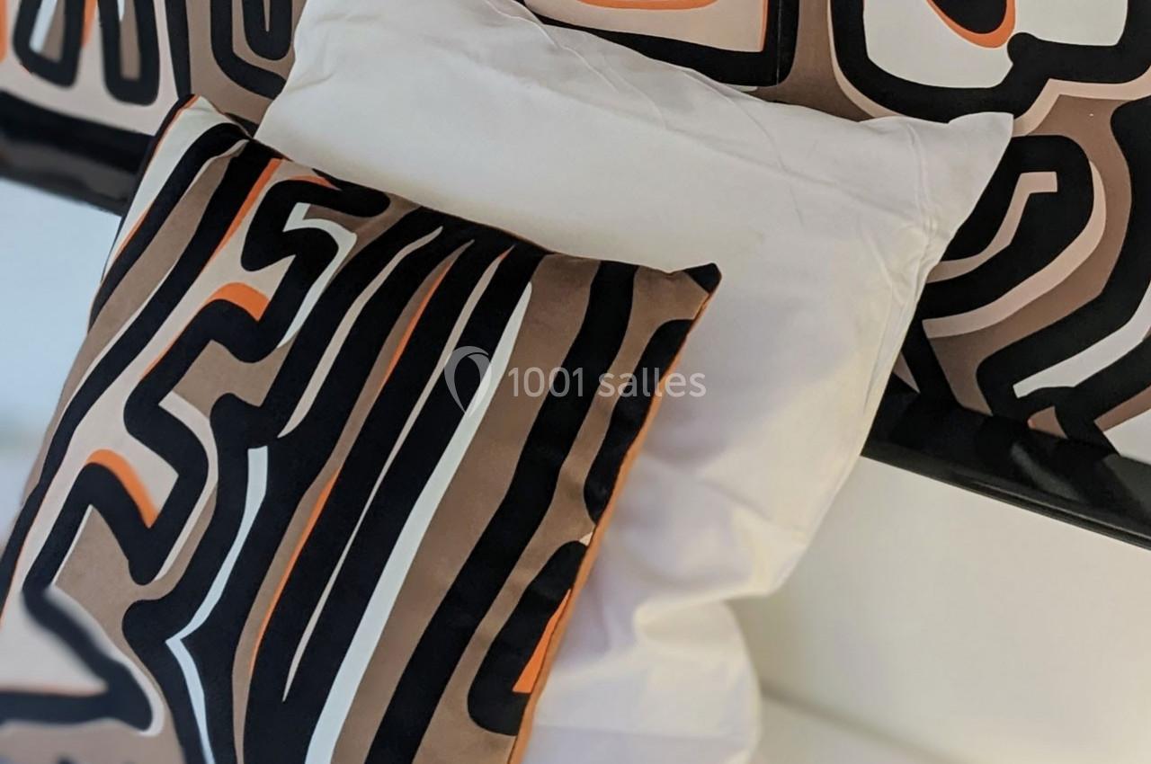 Coussins décoratifs avec motifs abstraits noirs, bruns et orange, posés sur un lit avec une housse blanche.