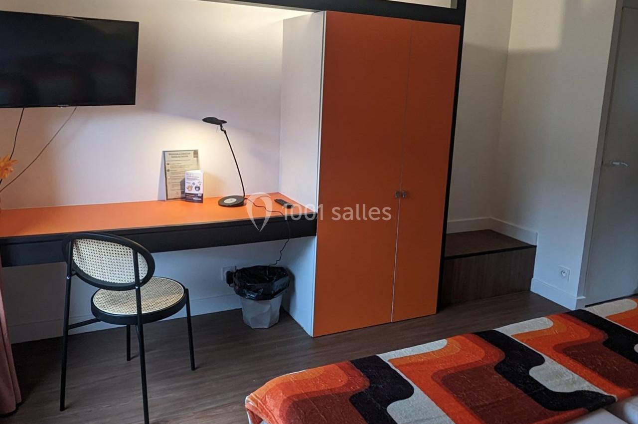 Bureau avec chaise, lampe, télévision murale et placard orange dans une chambre avec lit et couvre-lit coloré.