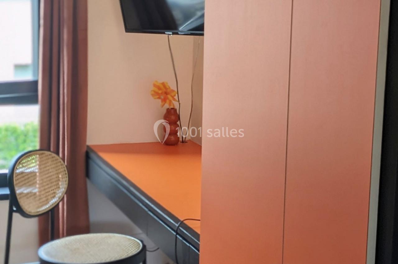 Bureau orange avec chaise en rotin, vase décoratif et télévision murale, près d'une fenêtre avec rideaux marron.