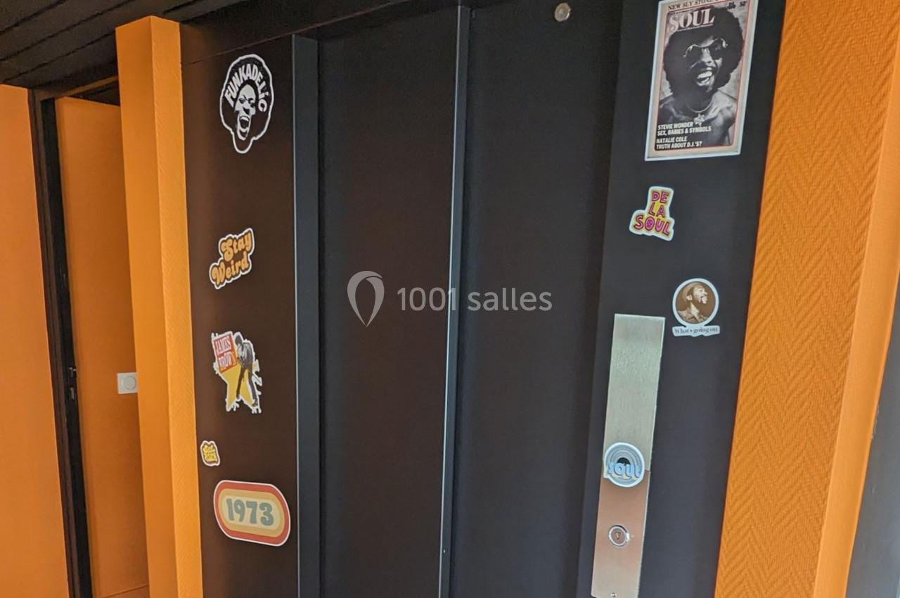 Porte d'ascenseur noire décorée d'autocollants colorés sur un mur orange vif.