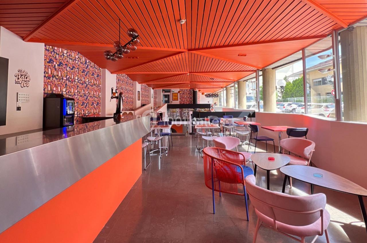 Salle lumineuse avec un bar moderne, des tables et des chaises colorées, et un plafond orange géométrique.