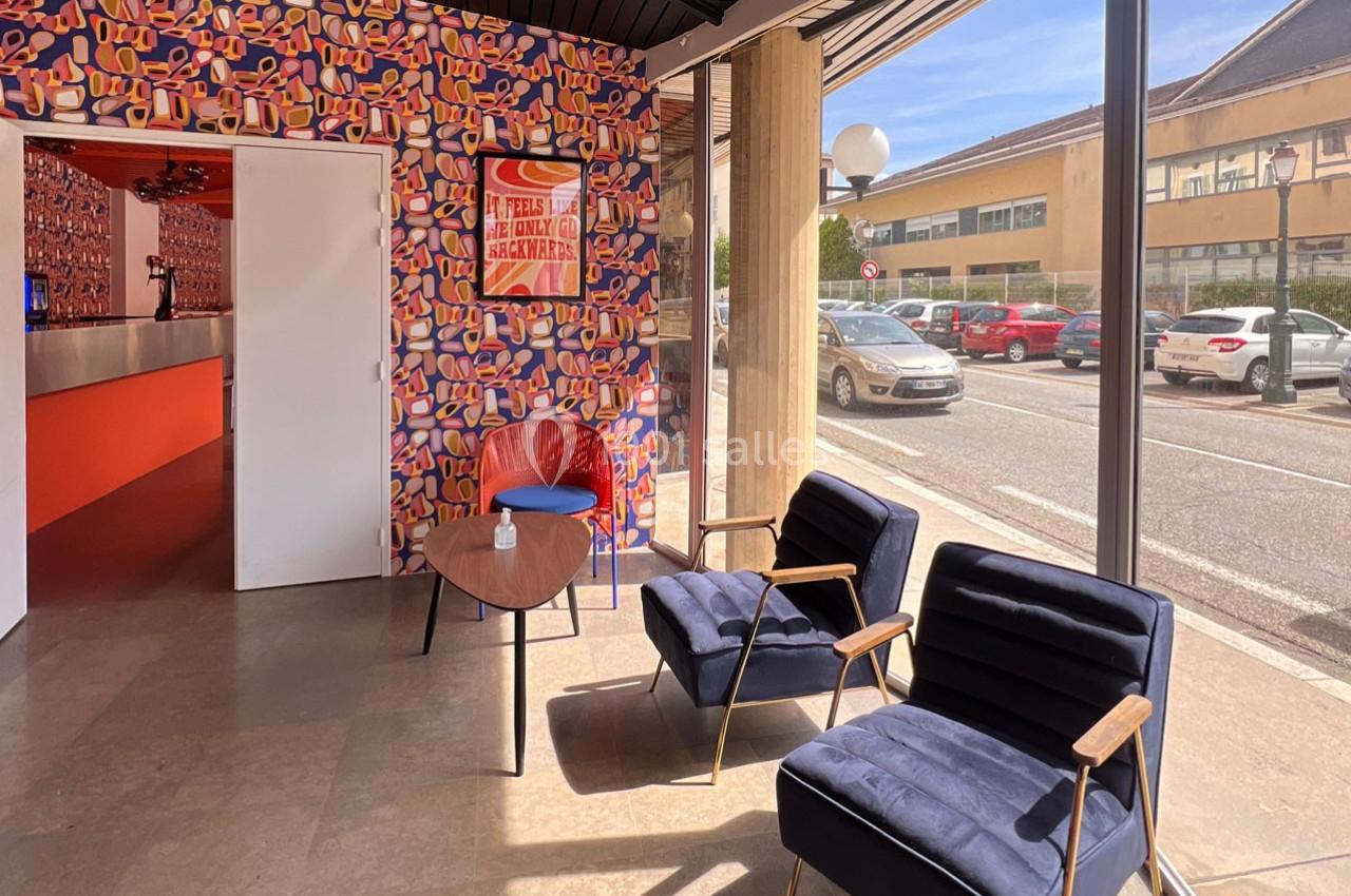 Espace d'attente lumineux avec fauteuils bleus, table basse en bois et mur coloré, vue sur la rue à travers une baie vitrée.