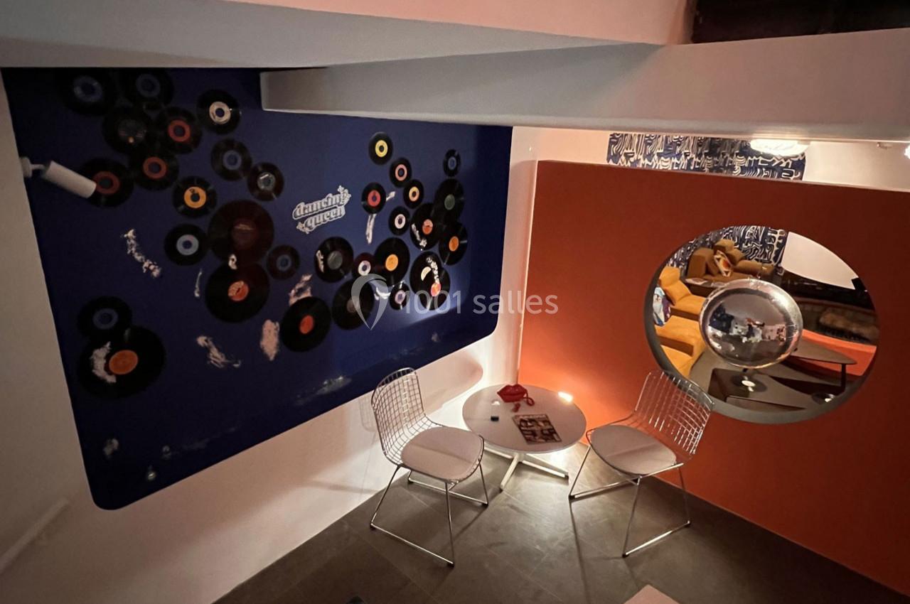 Mur décoré de vinyles, petite table ronde avec deux chaises blanches, miroir rond reflétant un canapé orange.