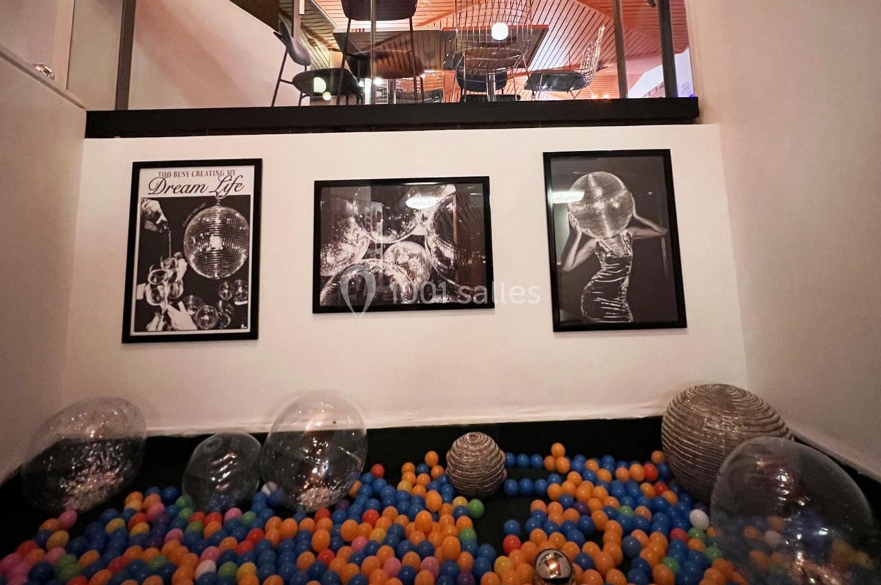 Piscine à balles colorées entourée de ballons transparents, avec des cadres photo en noir et blanc accrochés au mur.