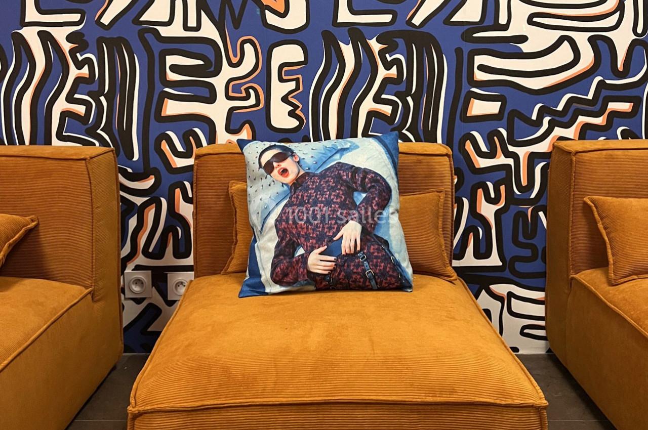 Fauteuil en velours orange avec un coussin imprimé d'un portrait, devant un mur décoré de motifs abstraits colorés.