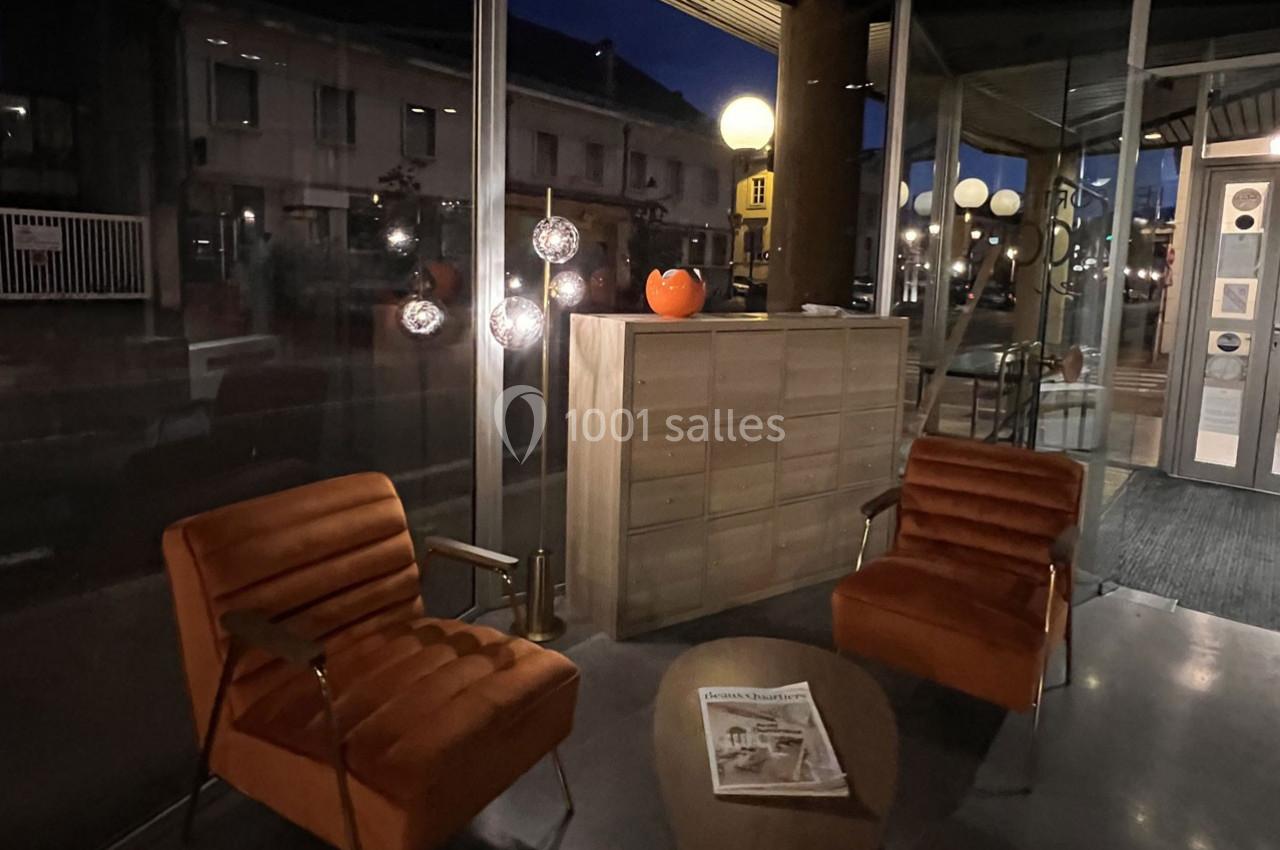 Deux fauteuils orange et une table basse avec un journal dans un hall vitré éclairé par des lampes en soirée.