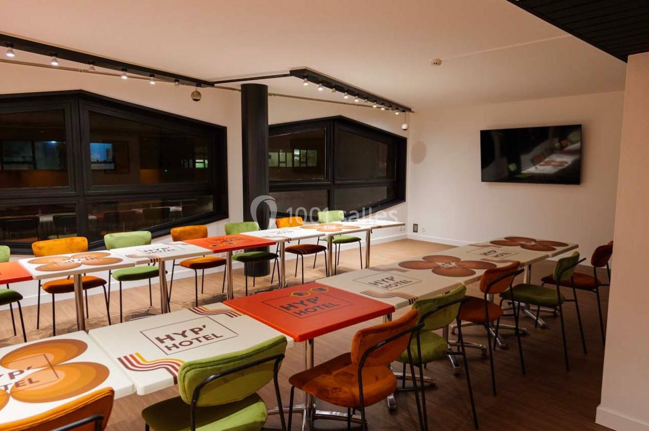 Salle de réunion lumineuse avec des tables colorées alignées, des chaises variées et un écran mural.