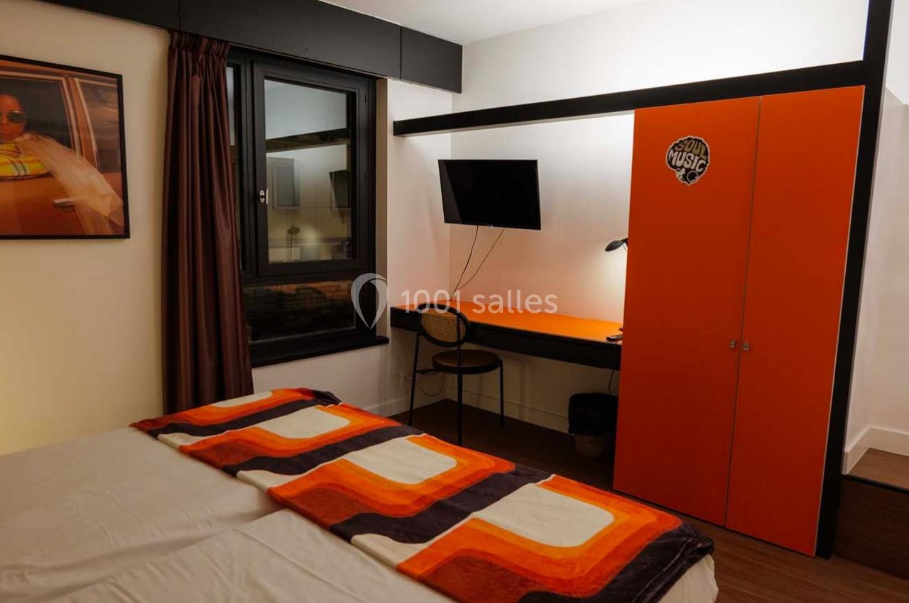 Chambre avec lit double, bureau orange, chaise, télé murale, armoire assortie et décoration murale.