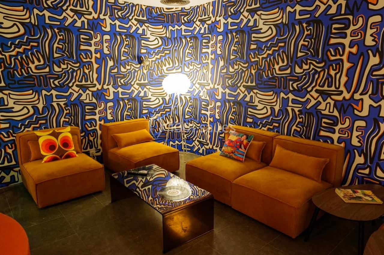 Salon coloré avec murs ornés de motifs abstraits bleus et noirs, mobilier jaune moutarde et éclairage central.