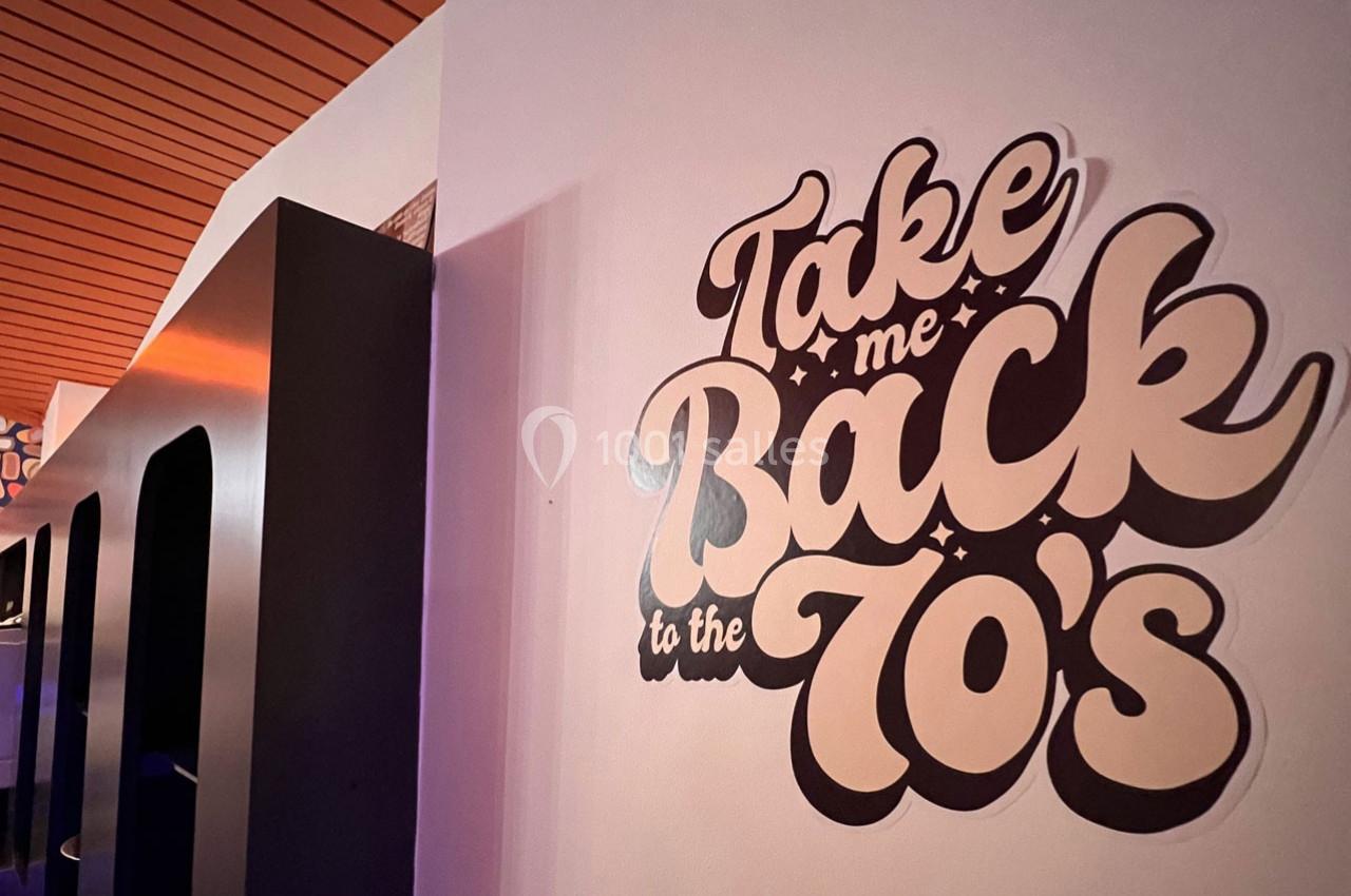 Texte ’Take me back to the 70's’ inscrit sur un mur blanc, avec un décor intérieur en arrière-plan.