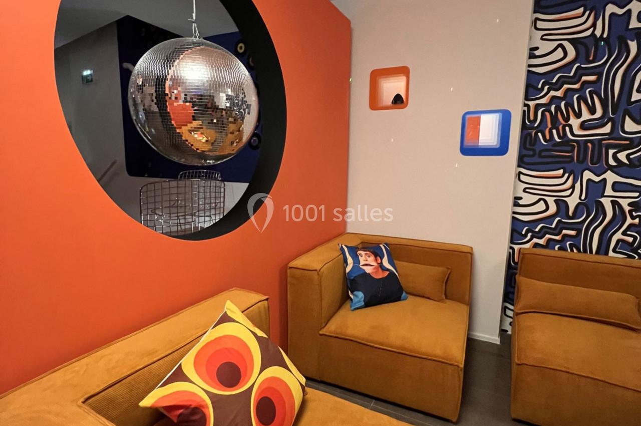 Salon moderne avec fauteuils en velours orange, coussins colorés, boule disco suspendue et décorations murales graphiques.