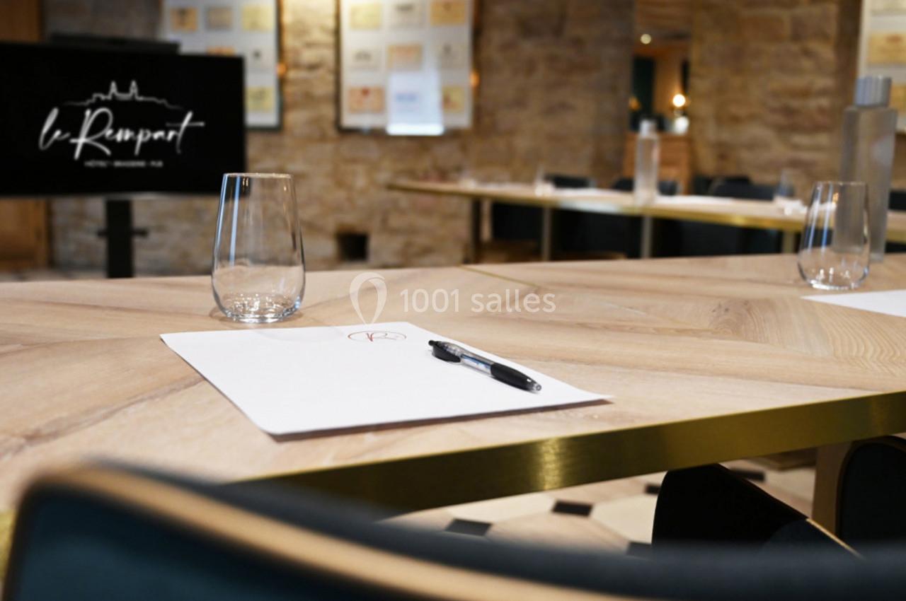 Salle de réunion avec table en bois, verres, feuilles et stylos, écran en arrière-plan et mur en pierre apparente.