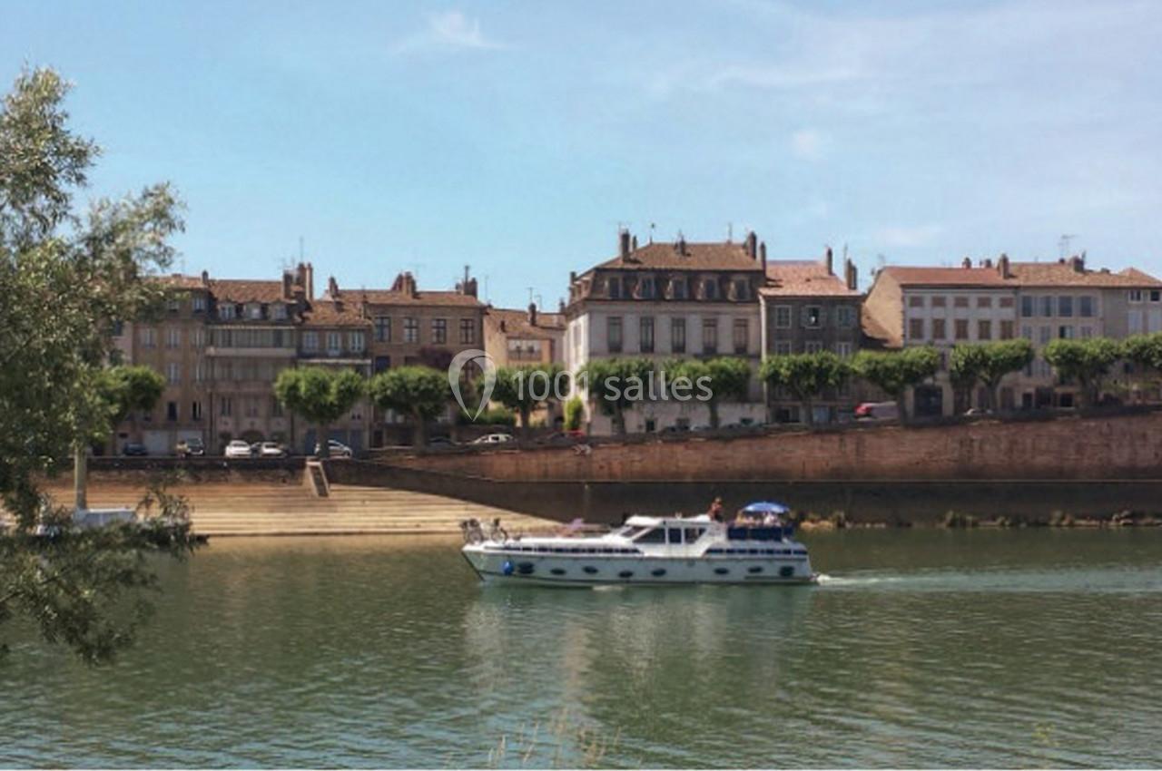 Bateau de plaisance naviguant sur une rivière bordée de maisons anciennes et d'arbres alignés.